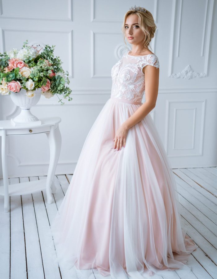 A beautiful bride with hair and makeup stands in a delicate pink wedding dress in a light decor with flowers
